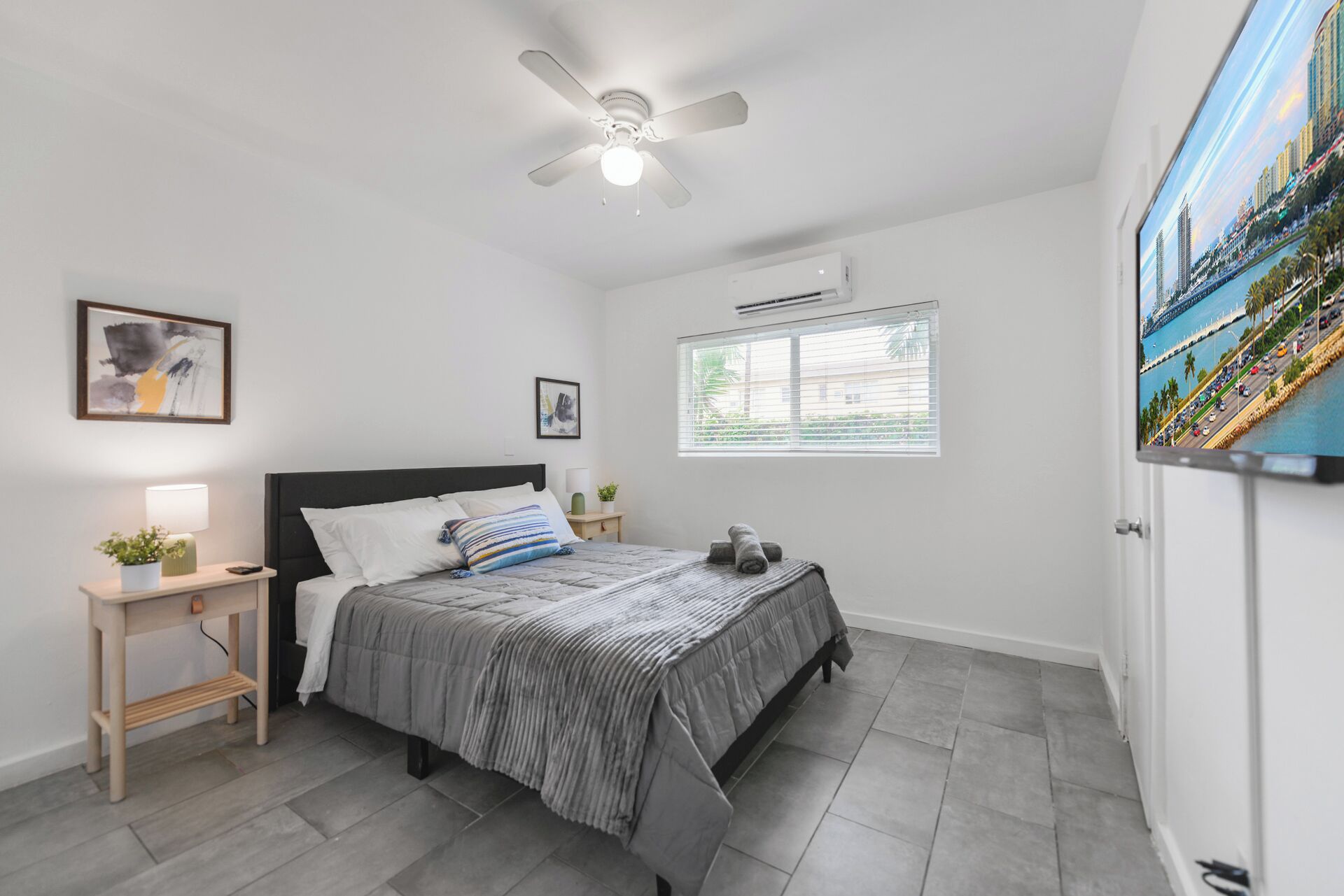 A bedroom with a bed, bedside tables, a ceiling fan, a window with blinds, and a cityscape painting on the wall.