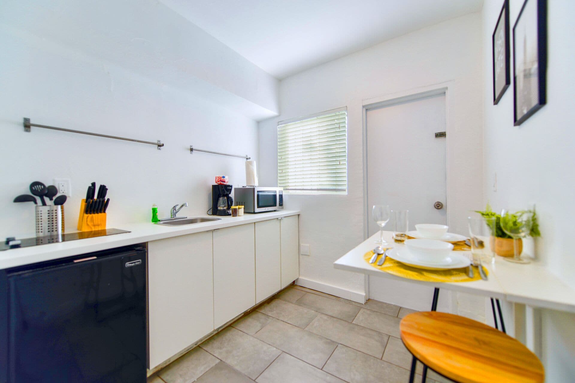 Une cuisine moderne avec des meubles blancs, un lave-vaisselle, un four à micro-ondes et une machine à café. On y trouve une fenêtre avec des stores, un évier et un petit coin repas avec une table et des chaises.