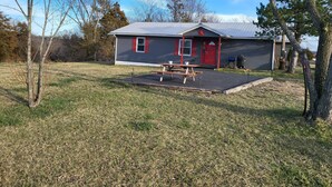 A small, single-story building with a red door and windows, a picnic table, and a barbecue grill outside. - The Shack- 3 bedroom finished house (Leadwood)
