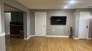 A spacious living room with hardwood flooring, a mounted flat-screen TV, and a door leading to a kitchen area. - The Shack- 3 bedroom finished house (Leadwood)