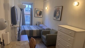 A bedroom with a bed, a chair, a dresser, a desk with a lamp, and two wall-mounted light fixtures. - Studio apartment in Original Brownstone in Harlem, New York (New York)