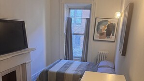 A small, cozy room with a bed, a wooden bench, a white table, a fireplace, a radiator, a window with curtains, and a wall-mounted TV.