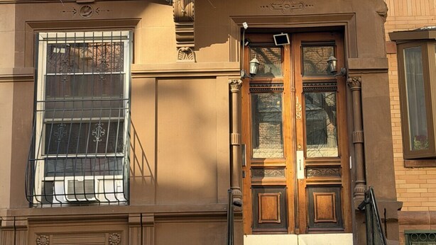 A brownstone building with a wooden front door, a window with a metal grill, and a visible address number.