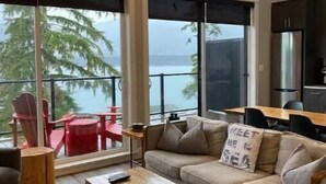 Een ruime woonkamer met een groot raam dat uitkijkt op een wateroppervlak, een houten salontafel en een bank met een kussen.
