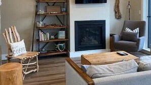 Een gezellige woonkamer met een open haard, een boekenkast, een comfortabele bank en een aan de muur bevestigde televisie.