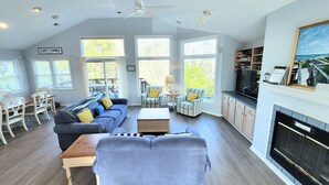 Un vaste salon comprenant un grand canapé gris, une table basse et des fauteuils à rayures. On y trouve également une cheminée, un téléviseur et un coin repas avec des chaises blanches et une table en bois.