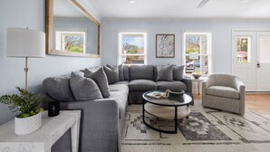 A modern living room with a grey sectional sofa, a round coffee table, and a small armchair. There is a ceiling fan, a floor lamp, and a large mirror on the wall. - Old Village - 3 BR with Private Backyard & Deck (Mount Pleasant)