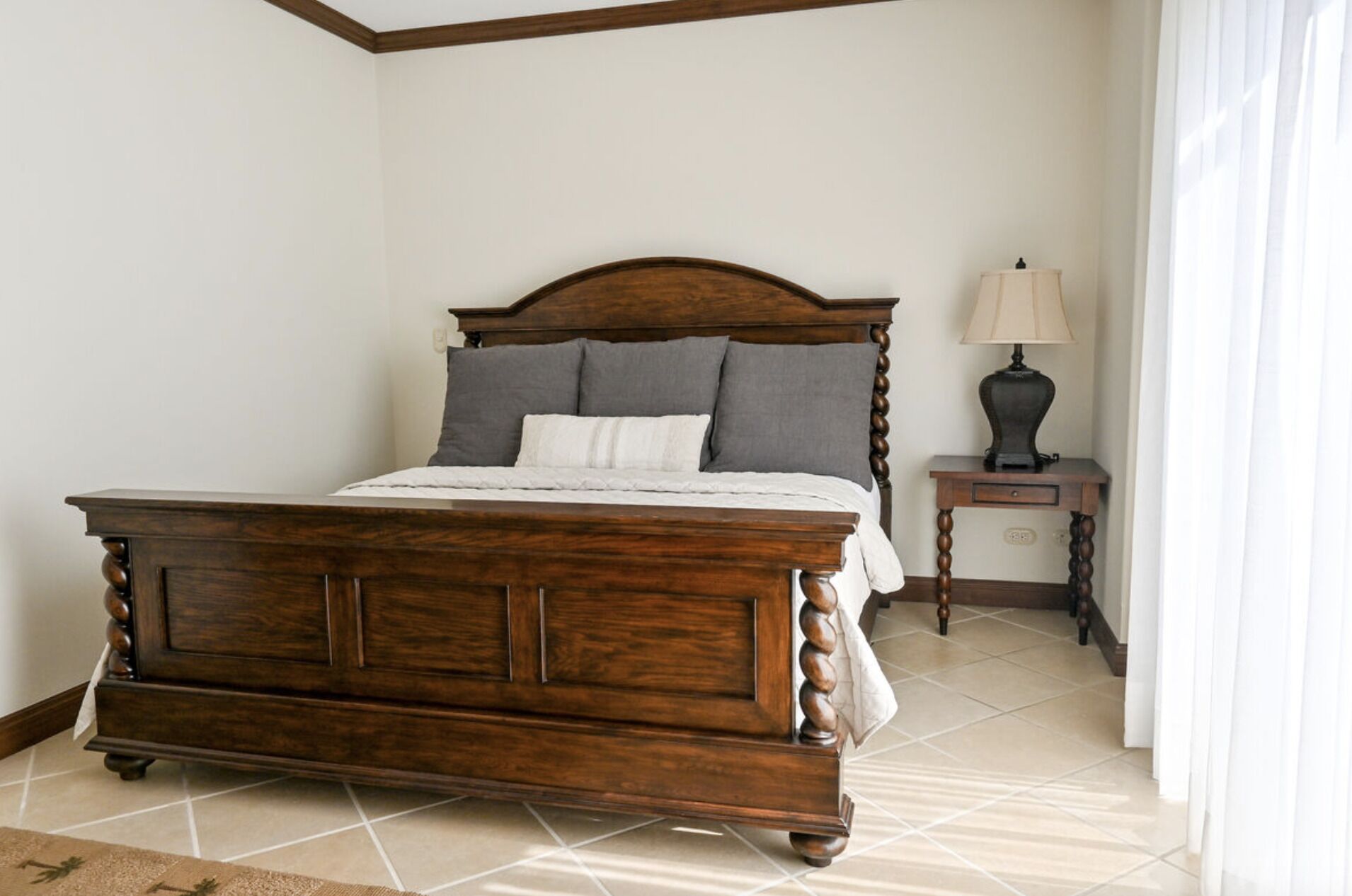 A wooden bed with a headboard, a bedside table with a lamp, and a tiled floor.