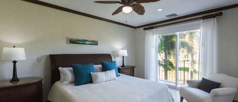 A bedroom with a large bed, two bedside tables with lamps, a ceiling fan, a window with curtains, and a view of the outdoors.