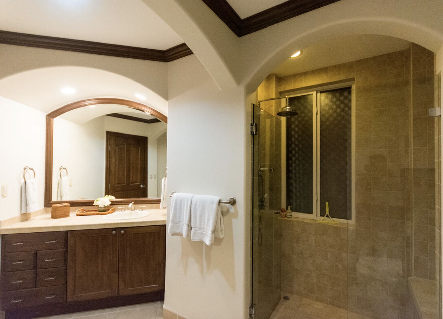Un baño con un espejo grande, muebles de madera, ducha con cerramiento de vidrio y tocador con lavamanos y toallero.
