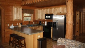 A wooden cabin interior with a kitchen featuring a central island, bar stools, a refrigerator, and a microwave. - Stunning Lake Petenwell Log Home Near Sand Valley Golf (Rome)