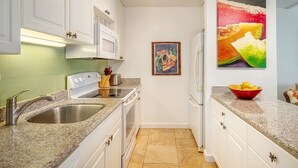 Eine Küche mit weißen Schränken, einer Granit Arbeitsplatte, einer Edelstahl-Spüle, einer Mikrowelle, einem Herd und einem Kühlschrank. An der Wand hängt ein gerahmtes Bild und auf der Arbeitsplatte steht eine Schüssel mit Obst.