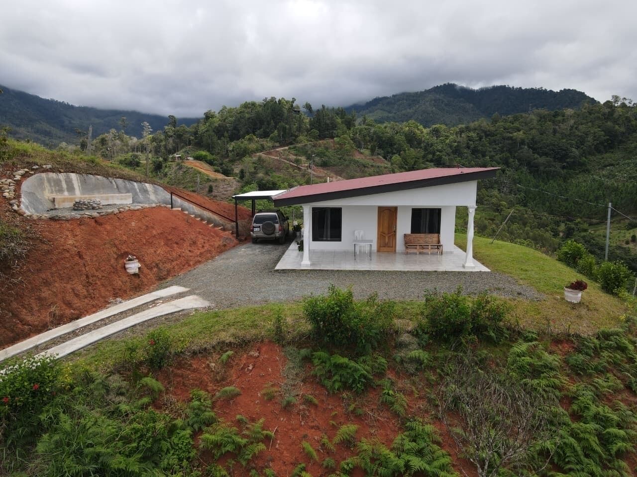 Una casa moderna con techo rojo, paredes blancas y un porche con asientos. Está rodeada por un terreno de tierra roja y vegetación verde.