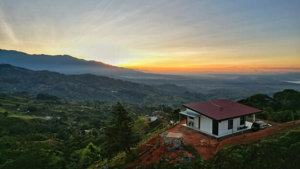 A modern house with a red roof, surrounded by lush green hills and mountains, during sunset. - A single-family home w/ a serene view. Nonsmoking and pet-friendly environment. (Perez Zeledon)