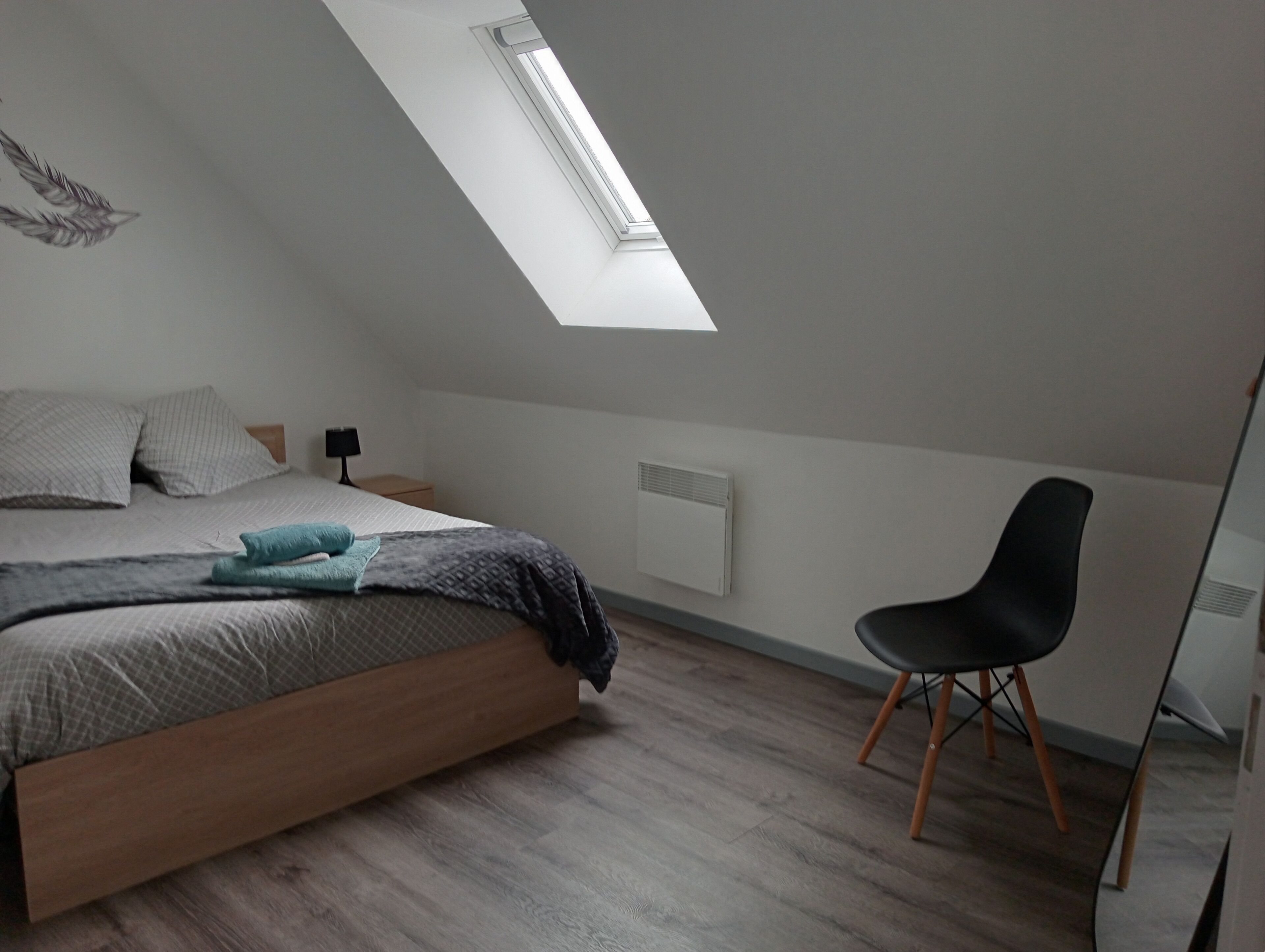 Une chambre avec un plafond en pente, un puits de lumière, un lit avec des oreillers, une table de chevet, une chaise et un radiateur.