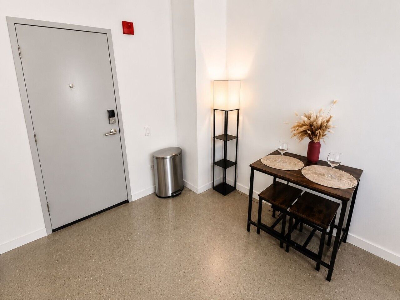 Una habitación con una puerta, un bote de basura, una mesa con dos sillas, una lámpara y un jarrón con flores secas.