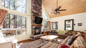 Un salón acogedor con chimenea de piedra, ventanas amplias, ventilador de techo y vistas al bosque.