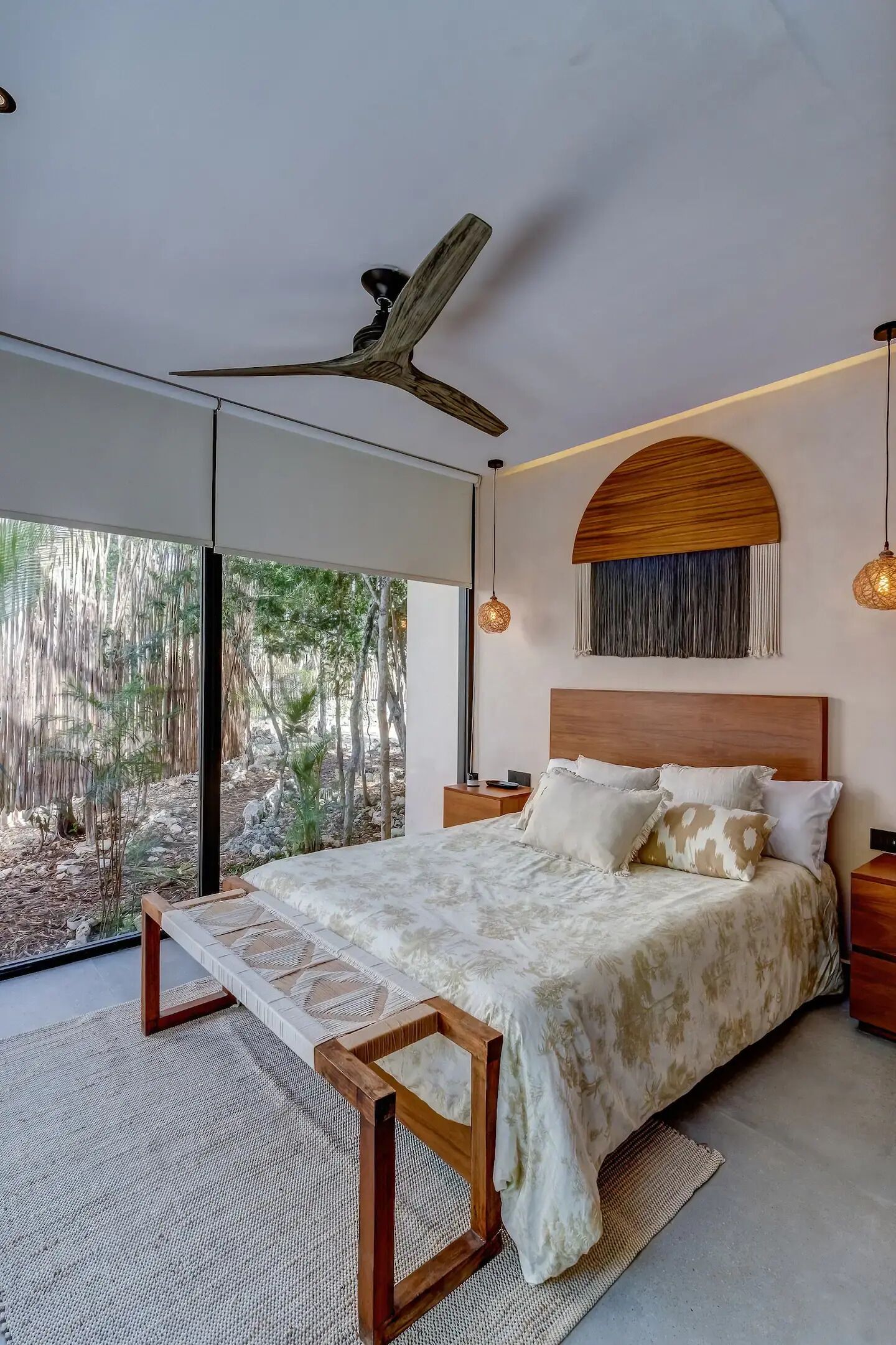 A modern bedroom with a large bed, a ceiling fan, and a view of a natural landscape through large windows.