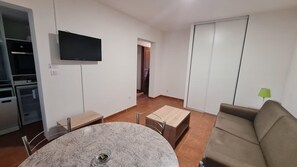 Un salon comprenant un téléviseur, un canapé, une table à manger avec des chaises, ainsi qu’un coin cuisine avec des placards et un four à micro-ondes.