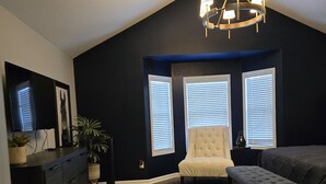 A room with a dark blue accent wall, a chandelier, a large window with blinds, a tufted armchair, a bench, a flat-screen TV, and a patterned rug.