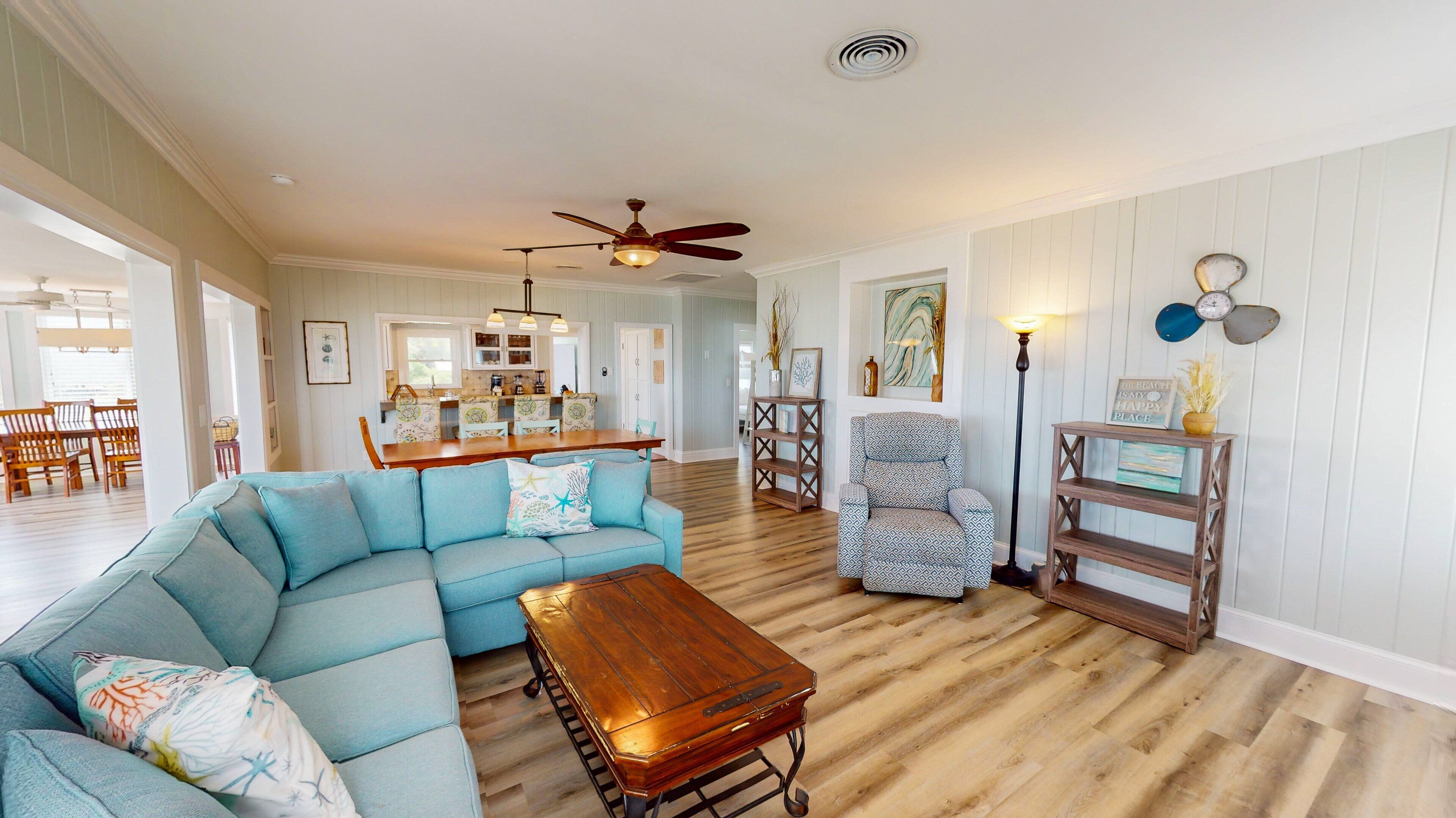 A spacious living room with a light blue sofa, a wooden coffee table, and a patterned armchair. A kitchen is visible in the background with wooden cabinets and a dining area.