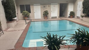 Una piscina interior rodeada de plantas en macetas, un suelo de hormigón y un edificio con múltiples puertas y ventanas al fondo.