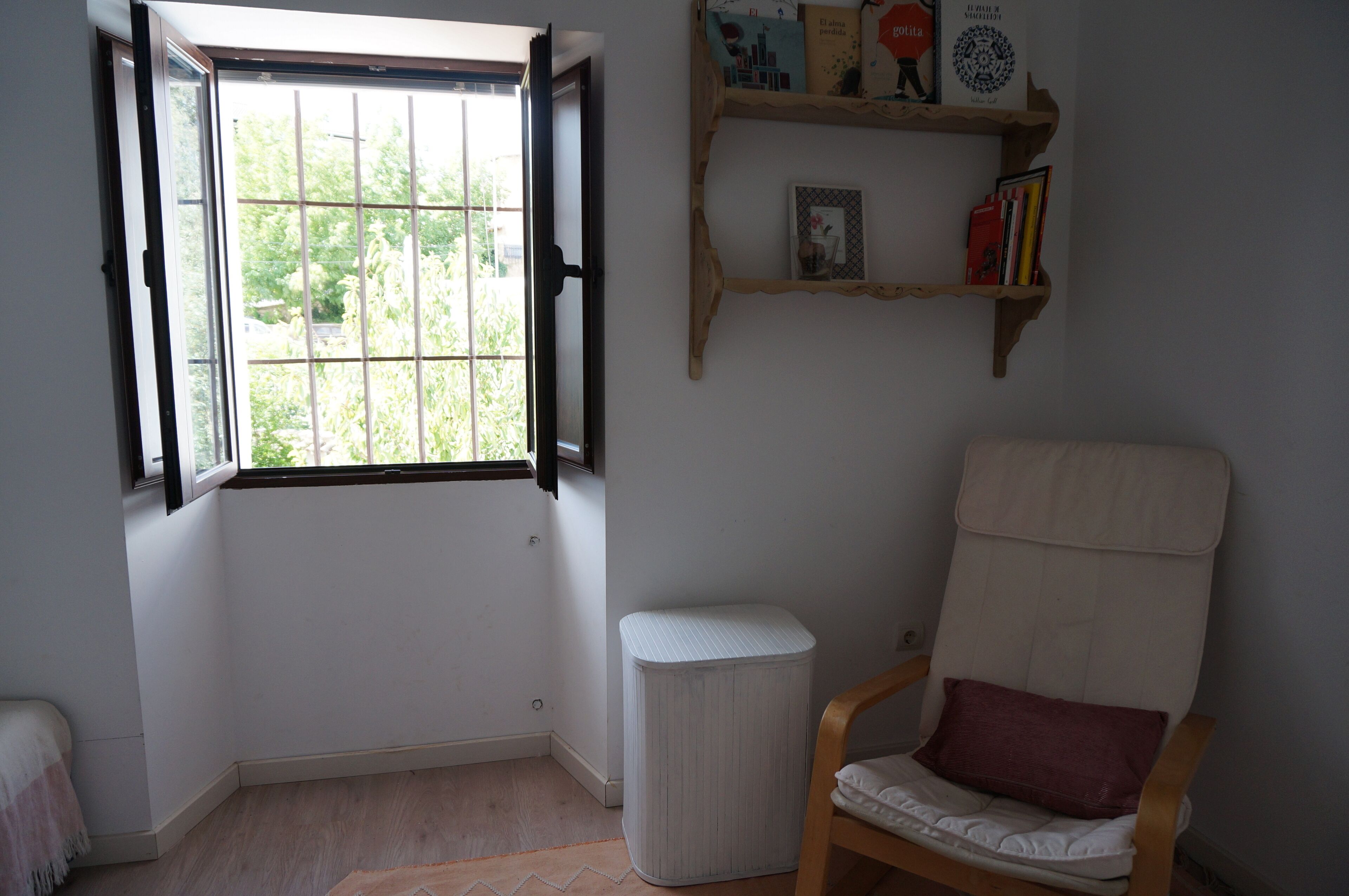 A room with a window, a chair, a small round table, and a shelf with books and decorations.
