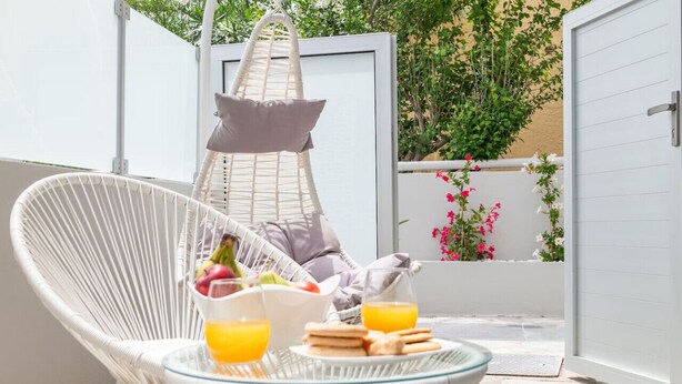 A wicker chair with a glass table, a hanging chair, and a door leading to a garden with flowers.