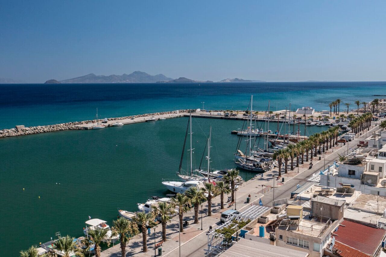 A marina with boats docked, a pier, and a coastal town with buildings and palm trees.
