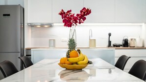 A modern kitchen with white cabinets, a stainless steel refrigerator, and a dining table with a fruit bowl. - Casa Lemonia - 110 sqm Maisonette (Kardamaina)