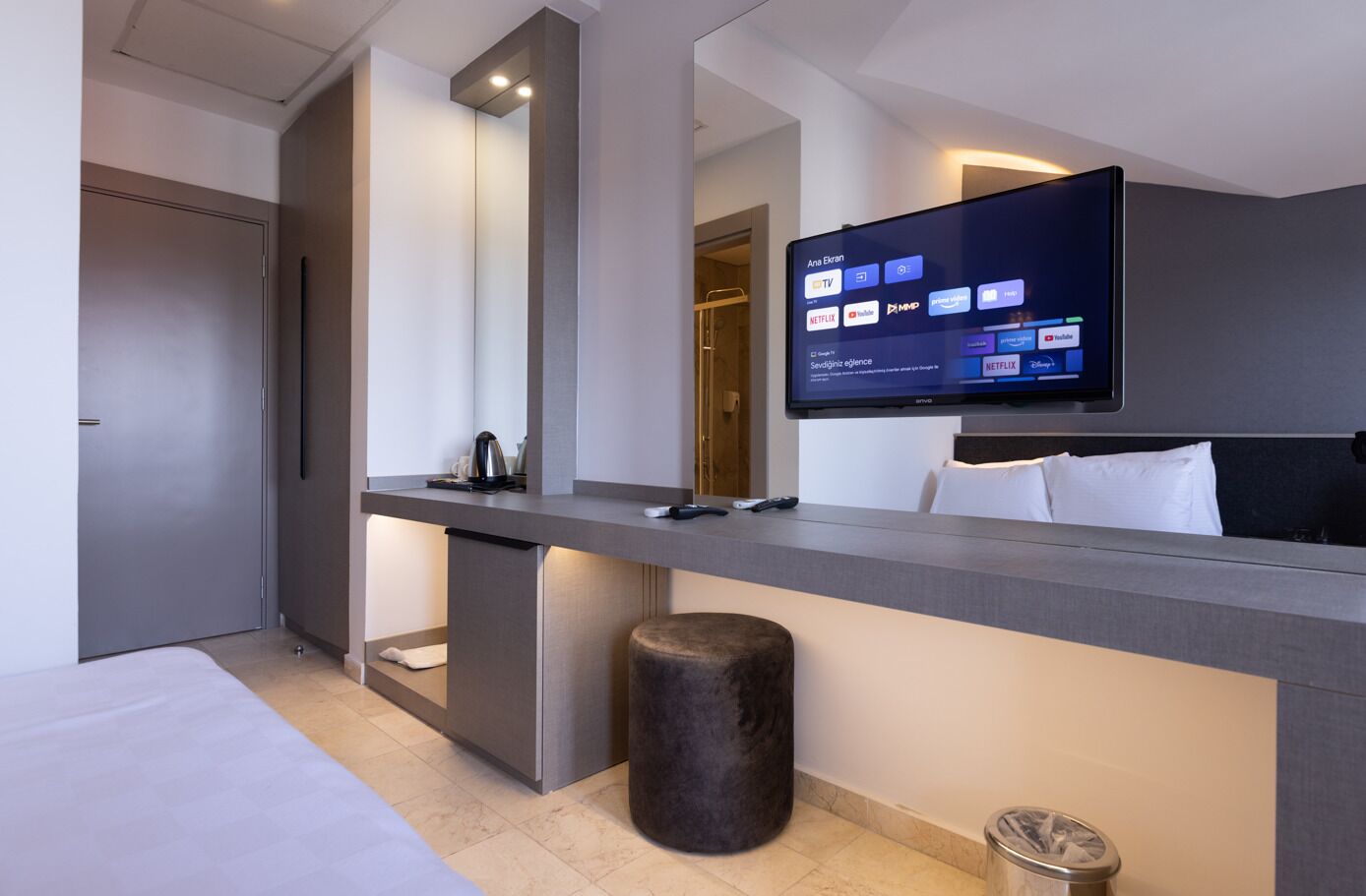 Ein modernes Hotelzimmer mit Fernseher, der Streaming-Dienste anzeigt, einem Bett, einem Schreibtisch mit Kaffeemaschine und einem Papierkorb.