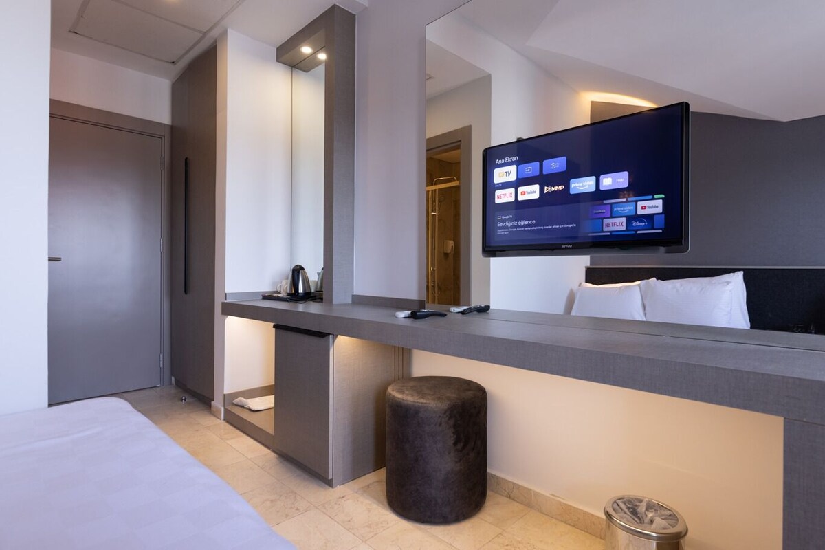 Ein modernes Hotelzimmer mit Fernseher, der Streaming-Dienste anzeigt, einem Bett, einem Schreibtisch mit Kaffeemaschine und einem Papierkorb.