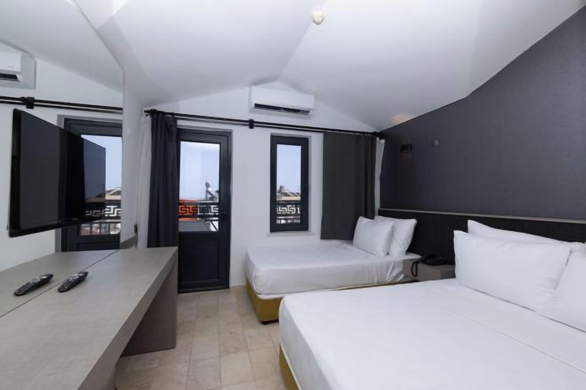 Ein Hotelzimmer mit einem Bett, einem Schreibtisch und einem Stuhl. Zwei Fenster mit Vorhängen und eine Wandklimaanlage sind vorhanden.