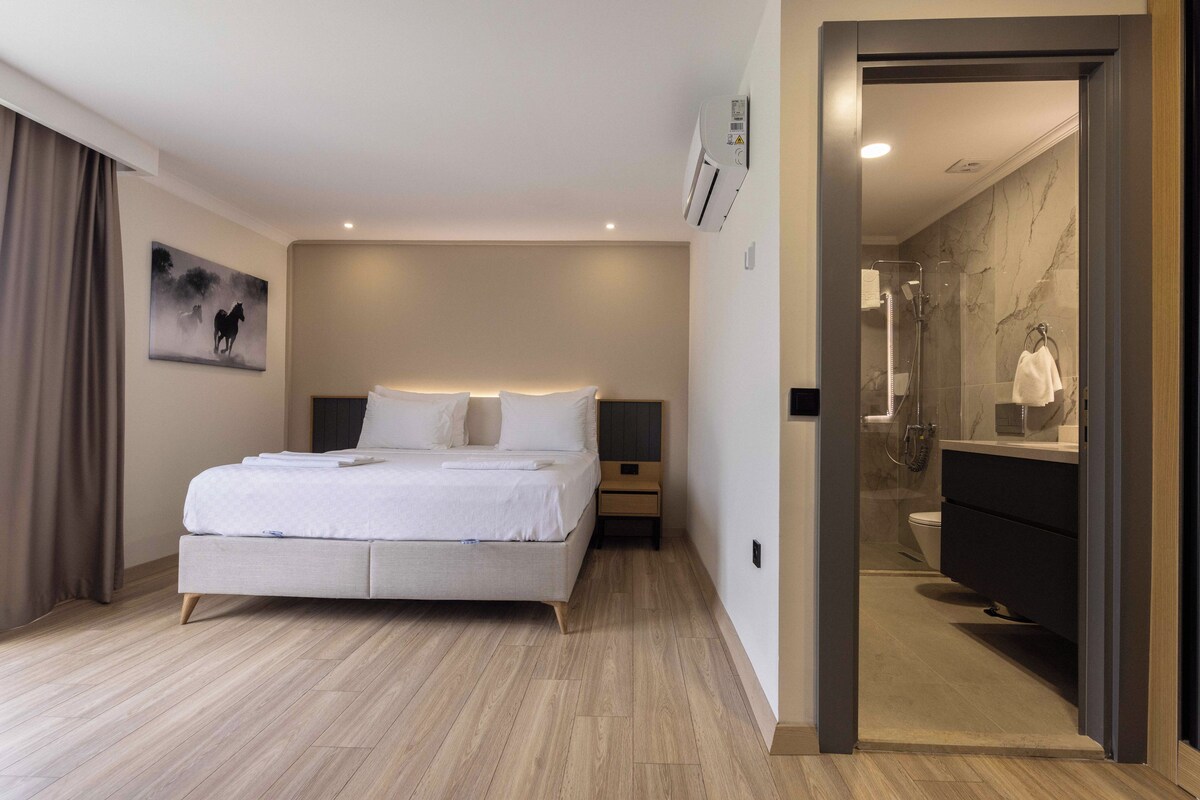 Ein modernes Hotelzimmer mit einem großen Bett, Nachttischen, einem Gemälde und einem Badezimmer mit Spiegel und Waschbecken.
