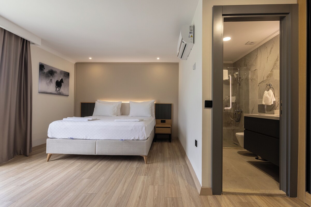 Ein modernes Hotelzimmer mit einem großen Bett, einem Nachttisch, einem Schreibtischchen, einem Bild an der Wand und einem Badezimmer mit Spiegel und Waschbecken.