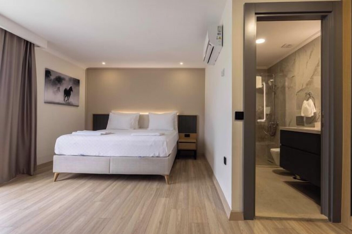 Ein modernes Hotelzimmer mit einem großen Bett, einem Nachttisch, einem Schranken und einem an der Wand hängenden Gemälde. Ein Badezimmer ist durch eine offene Tür sichtbar.