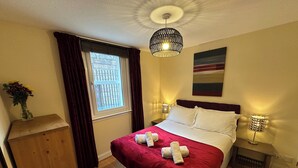 A neatly arranged bedroom with a large bed, two bedside lamps, a window with curtains, and a hanging light fixture. - 2 bedroom property with a fantastic view of Edinburgh Castle in the heart of the Old Town (Edinburgh)