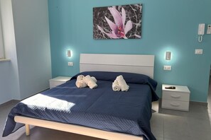 Ein ordentlich bezogenes Bett mit zwei zusammengerollten Handtüchern, ein weißer Nachttisch mit einem Telefon und eine Wandleuchte.