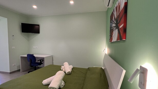 Quarto de hotel com cama, escrivaninha, cadeira e televisão na parede.