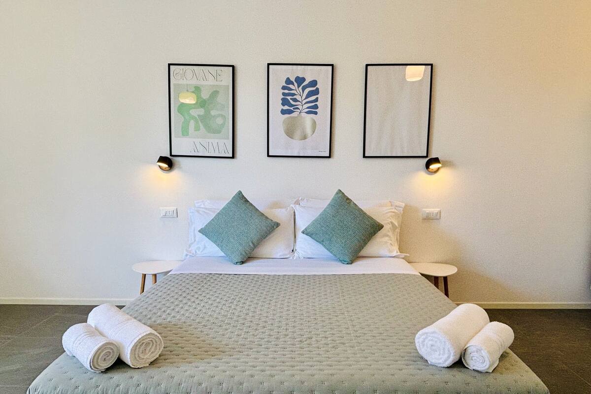 A neatly made bed with two pillows, a green blanket, and rolled towels. There are three framed artworks on the wall above the bed.