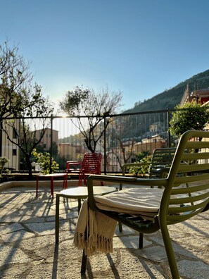 A terrace with a green metal chair and a red chair, both with cushions. The terrace has a stone floor and is surrounded by a metal fence. - Villa Reise (Riomaggiore)