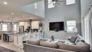 A modern living room with a sofa, a coffee table, and a TV mounted on the wall. There is a kitchen area in the background with white cabinets and a dining table. - Dream getaway just 2 miles from Fenwick or OC beach.  5BR house sleeps up to 13 (Selbyville)