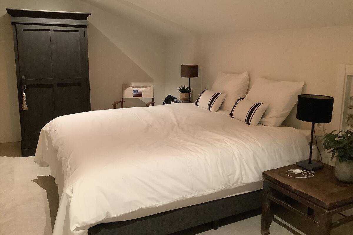 Een slaapkamer met een groot bed, een houten garderobe, een nachtkastje met een lamp en een kleine plant.
