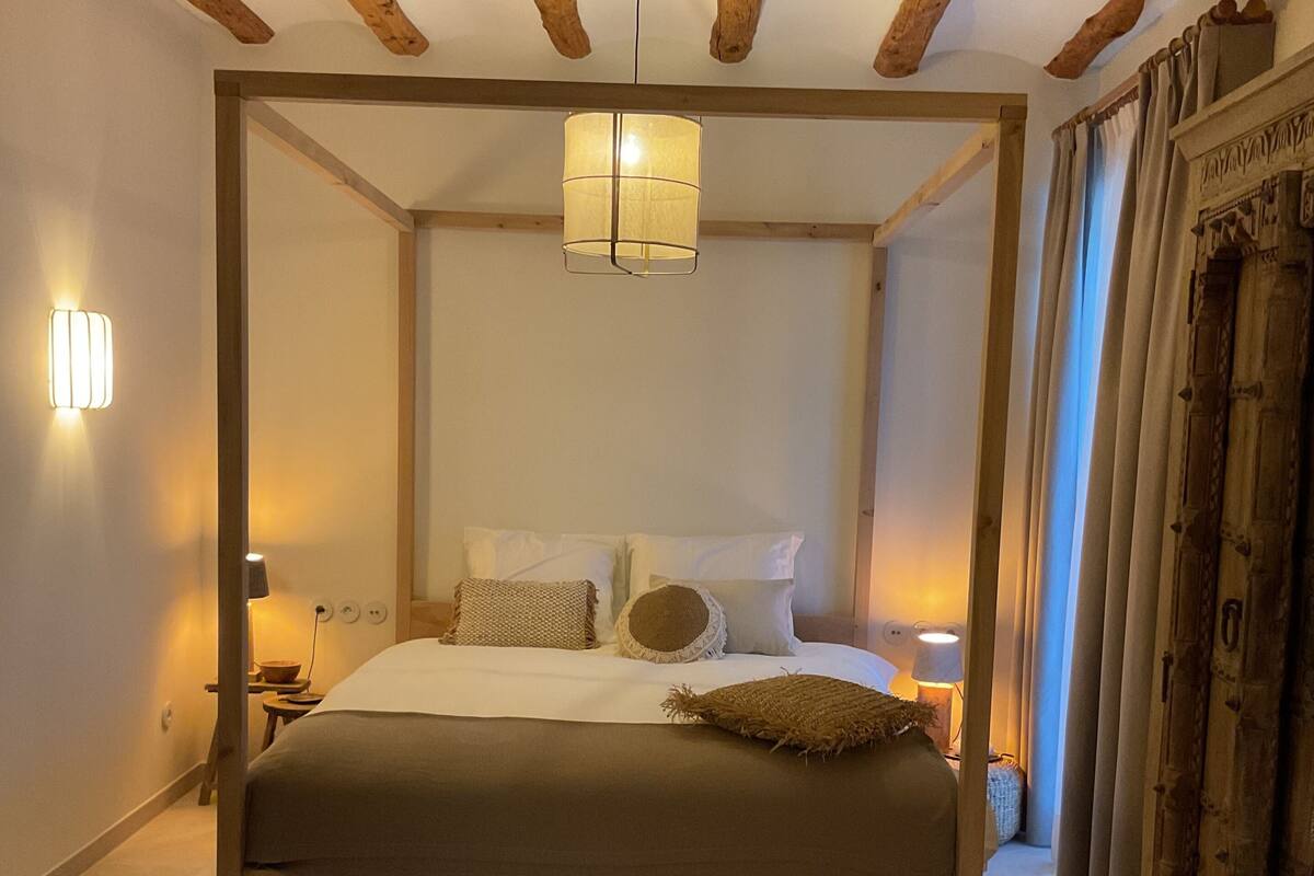 Een hemelbed met houten frame, een hanglamp en een nachtkastje met lamp.