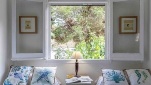 Un dormitorio con dos camas, una ventana con vista a zonas verdes, una mesita de noche con lámpara y cuadros en las paredes.