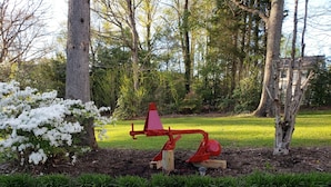 Un parque infantil con un tobogán rojo y un balancín pequeño, rodeado de árboles y césped.