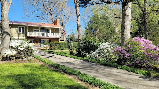 Una casa de dos plantas con un porche delantero, rodeada de jardines cuidados y arbustos florecientes.