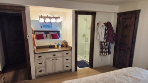 A bedroom with a large bed, a bathroom with a sink, mirror, and shower, and a wooden door.