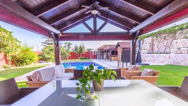 Un patio al aire libre con una mesa de vidrio, sillas de mimbre, una planta en maceta y una vista de una piscina y una casa al fondo.