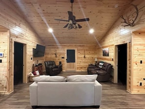 Ein Interieur einer Blockhütte mit einem weißen Sofa, zwei Ledersesseln, einem Deckenventilator und einem Wand-Fernseher.